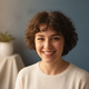 Smiling young woman with short curly dark hair in a cream top, looking at the camera. Soft slate blue background.